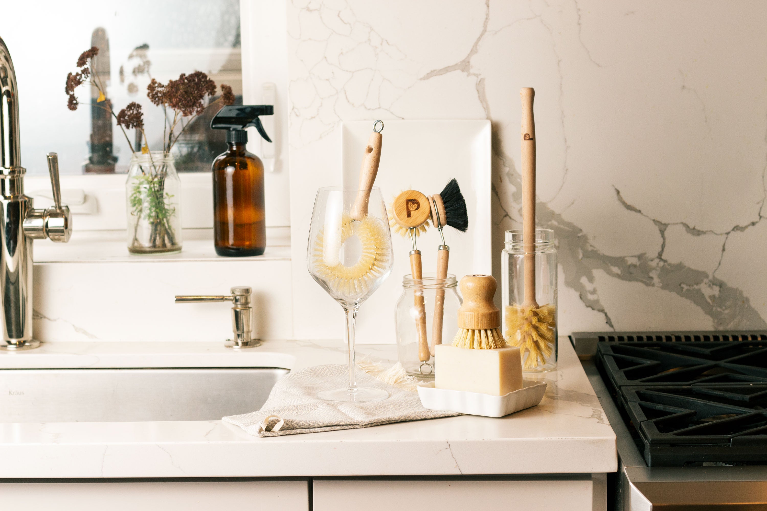 Eco-friendly wooden dish brushes, soap bar, amber spray bottle, and wine glass on a white marble kitchen counter near a sink.