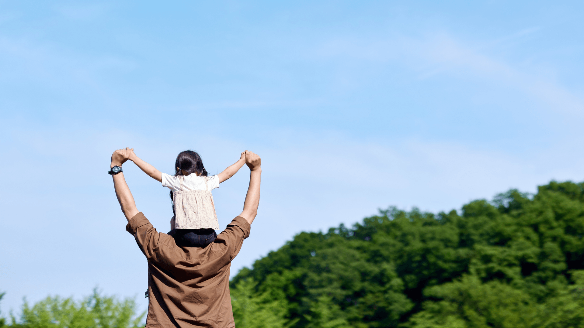 Man with child on shoulders against a blue sky with trees below